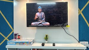 Un televisor de pantalla plana montado en un estante blanco sobre una pared azul con motivos geométricos amarillos.