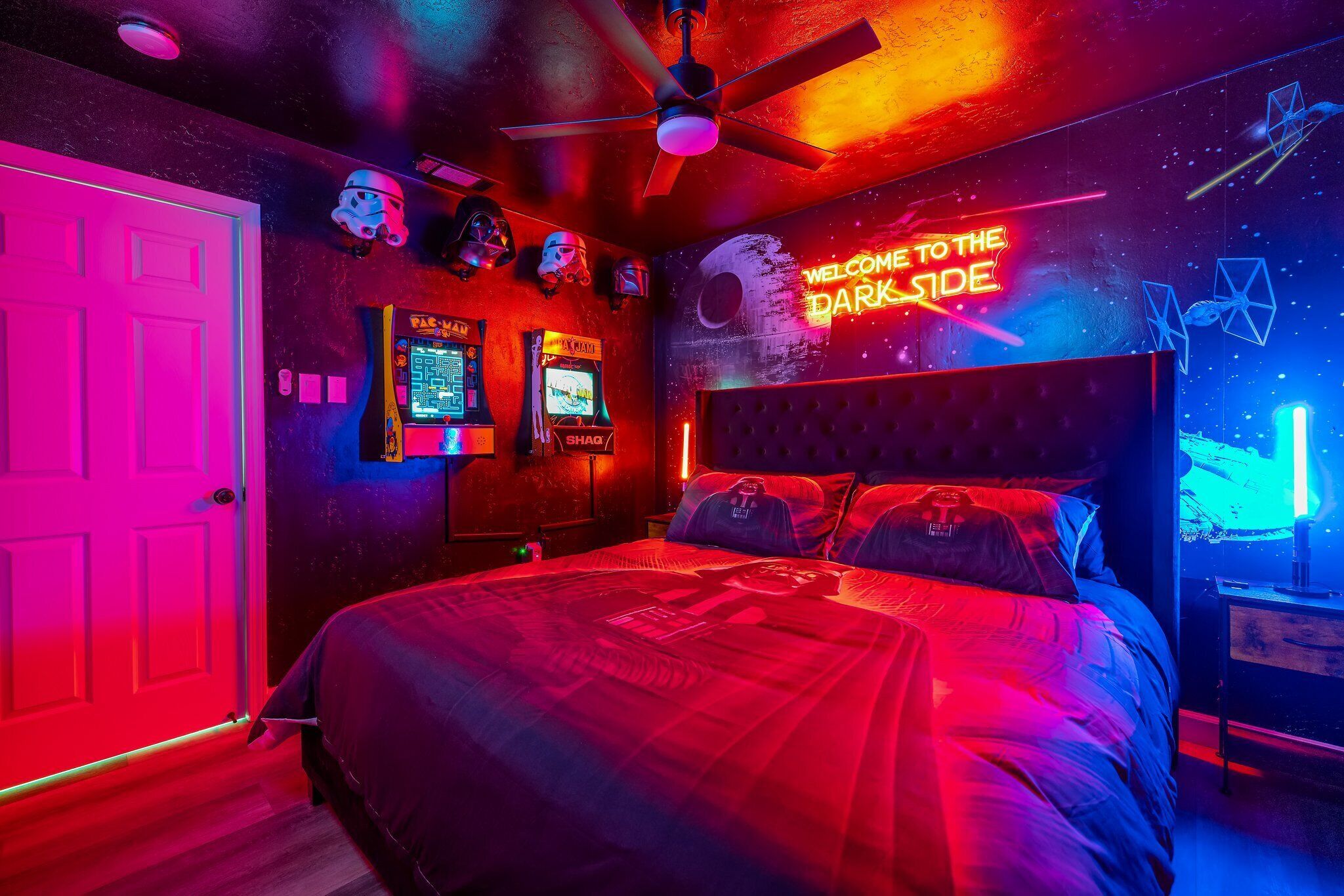 A room with a bed, ceiling fan, neon lights, and arcade machines.