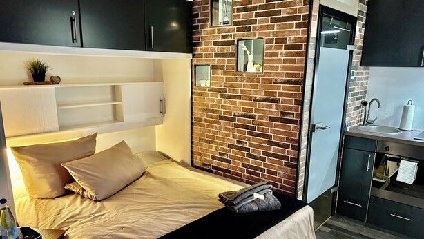 Une chambre moderne avec un mur en briques, un lit avec une literie beige, une table de chevet avec une bouteille et une petite plante.
