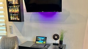 Un salon moderne avec un téléviseur à écran plat fixé au mur, un ordinateur portable posé sur une table noire, deux fauteuils gris, une petite plante en pot et un distributeur automatique de collations.
