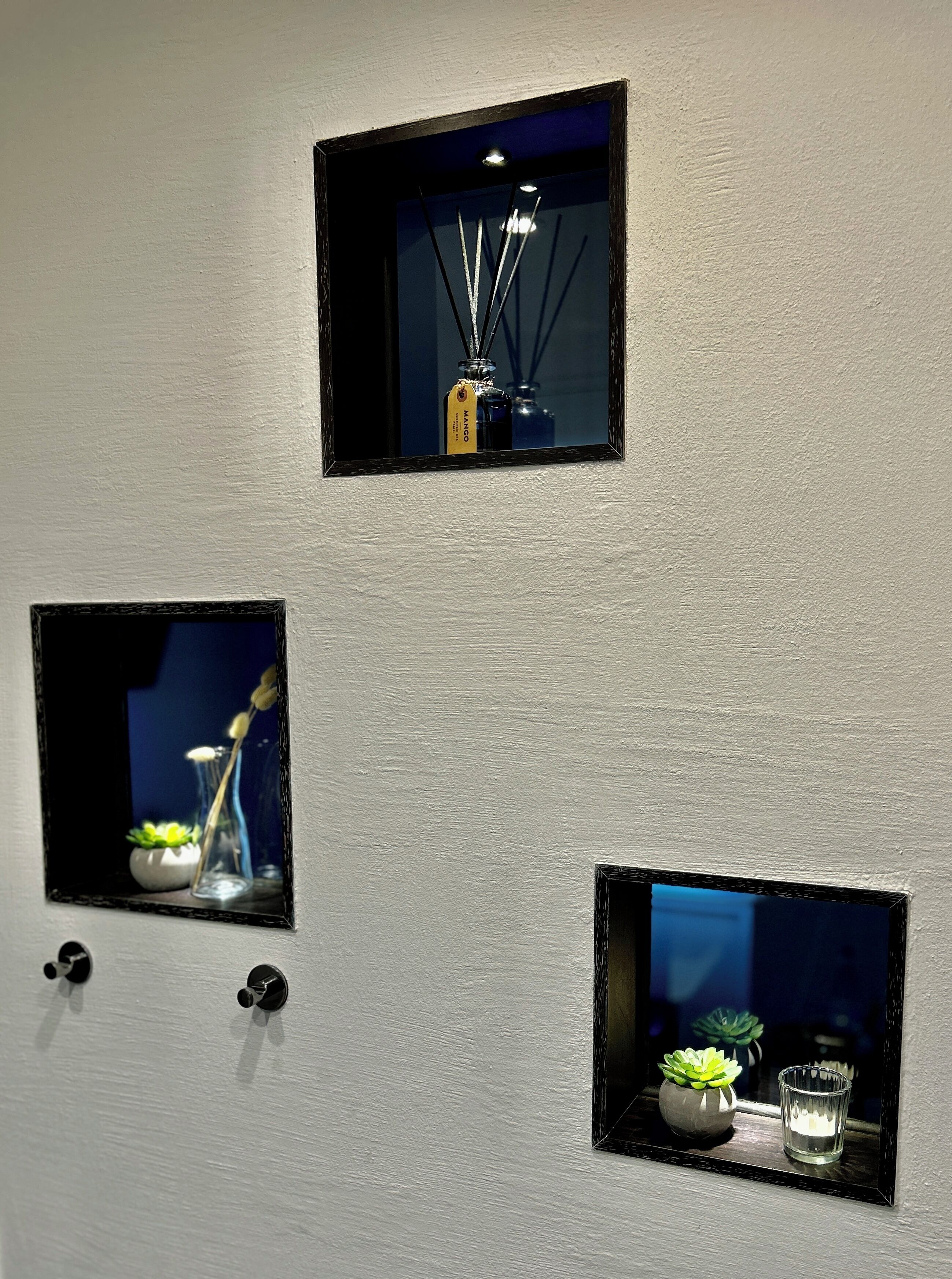 Drei quadratische Spiegel an einer Wand, jeder mit einem anderen Dekorationselement: eine Flasche, ein Glas mit Pflanze und eine Kerze.