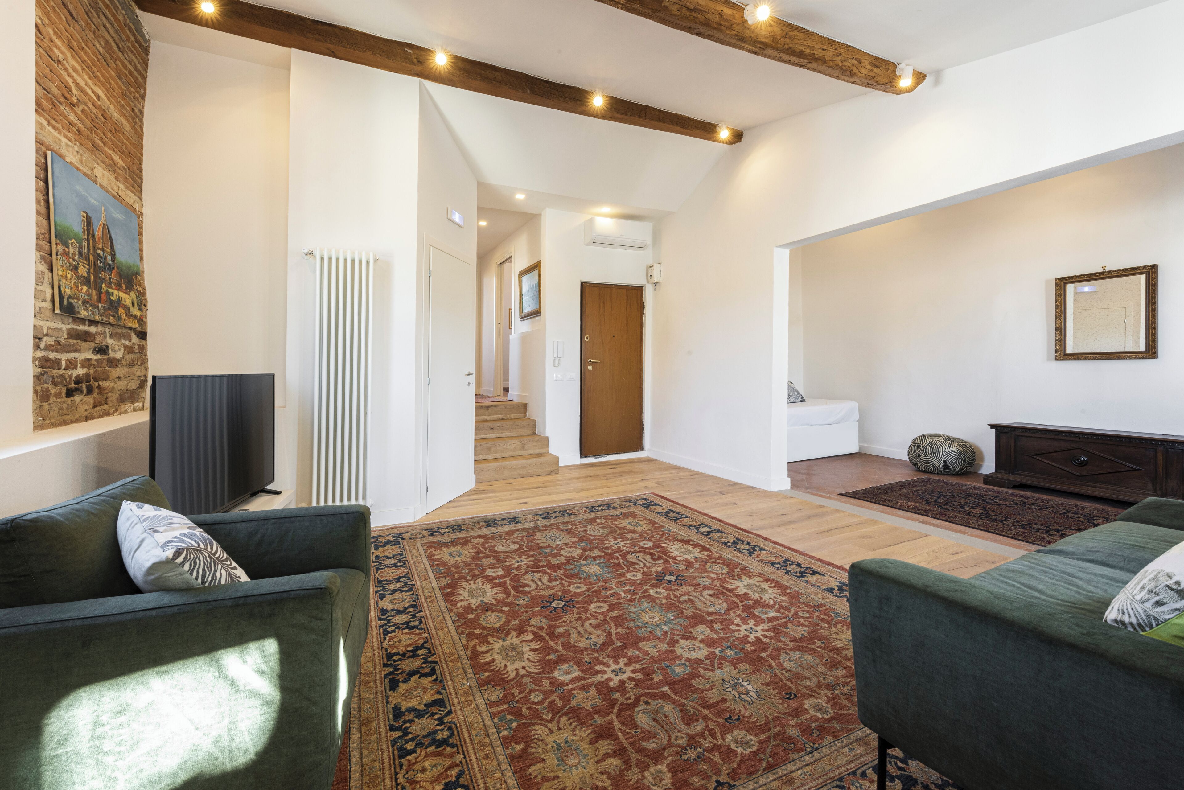 A spacious living area with a large rug, two green sofas, a television, and a staircase leading to another room.
