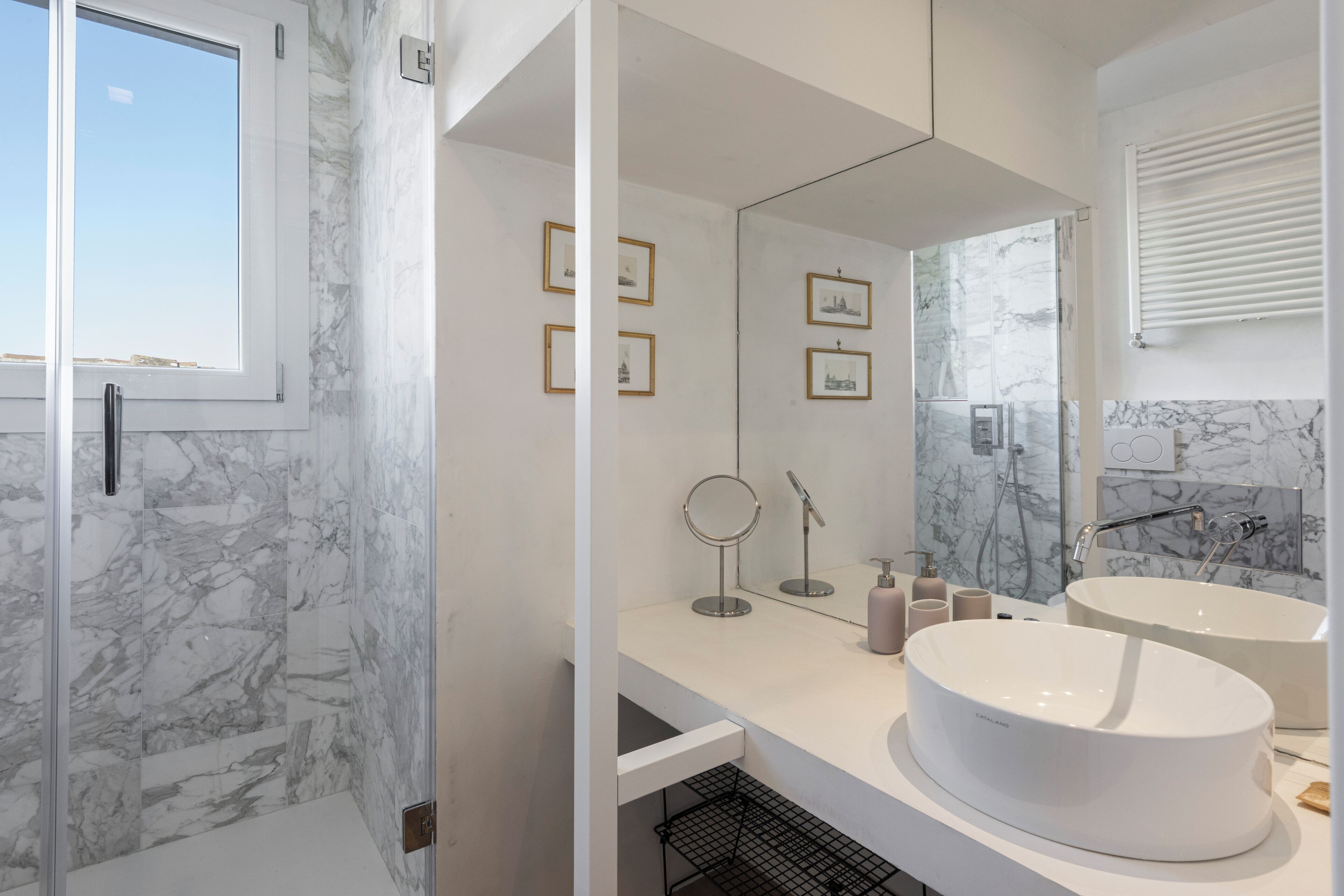 A modern bathroom with marble walls, two large sinks, a glass shower enclosure, and framed pictures on the wall.