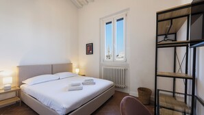 Ein Schlafzimmer mit Bett, Nachttischlampen, einem Fenster, einer Heizung, einem Korbwägelchen und einem Sessel.