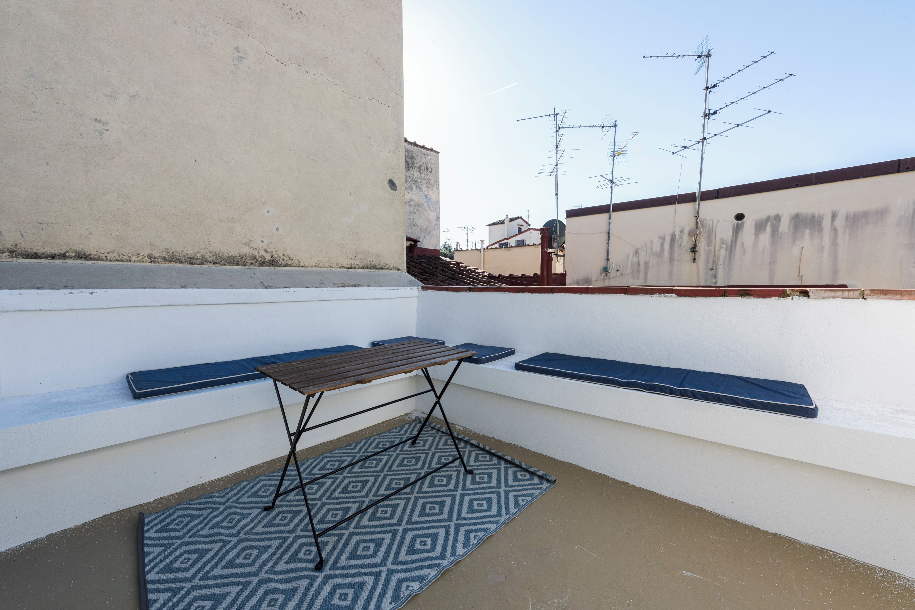 A rooftop terrace with a table and benches, a patterned rug, and a view of neighboring buildings.
