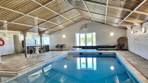 Una piscina interior con suelo de baldosas, techo metálico y una zona de estar con mesas y sillas.
