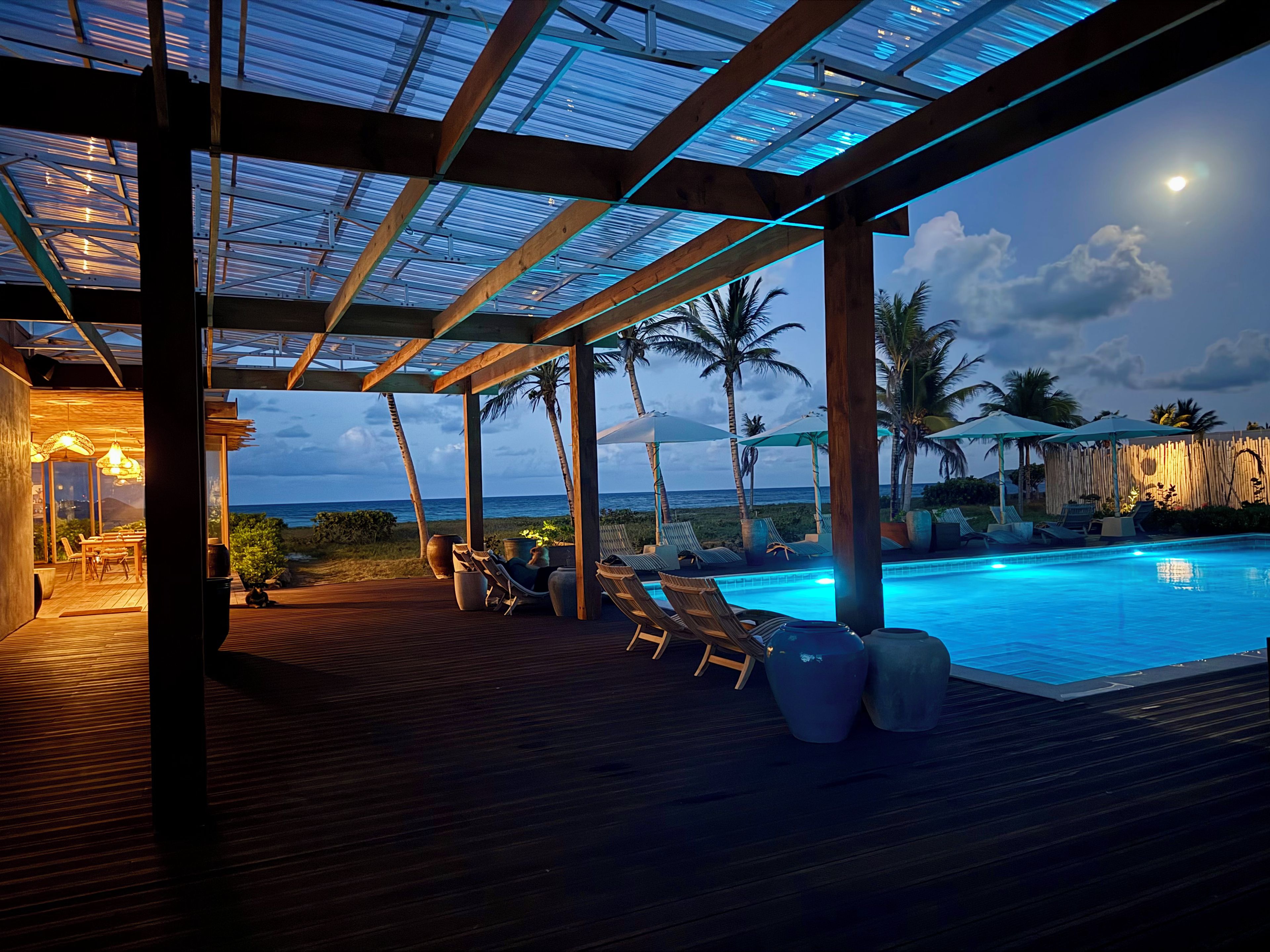 An outdoor pool area with lounge chairs and umbrellas, wooden deck flooring, and a view of the ocean.