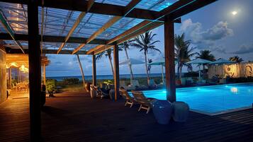 An outdoor pool area with lounge chairs and umbrellas, wooden deck flooring, and a view of the ocean.