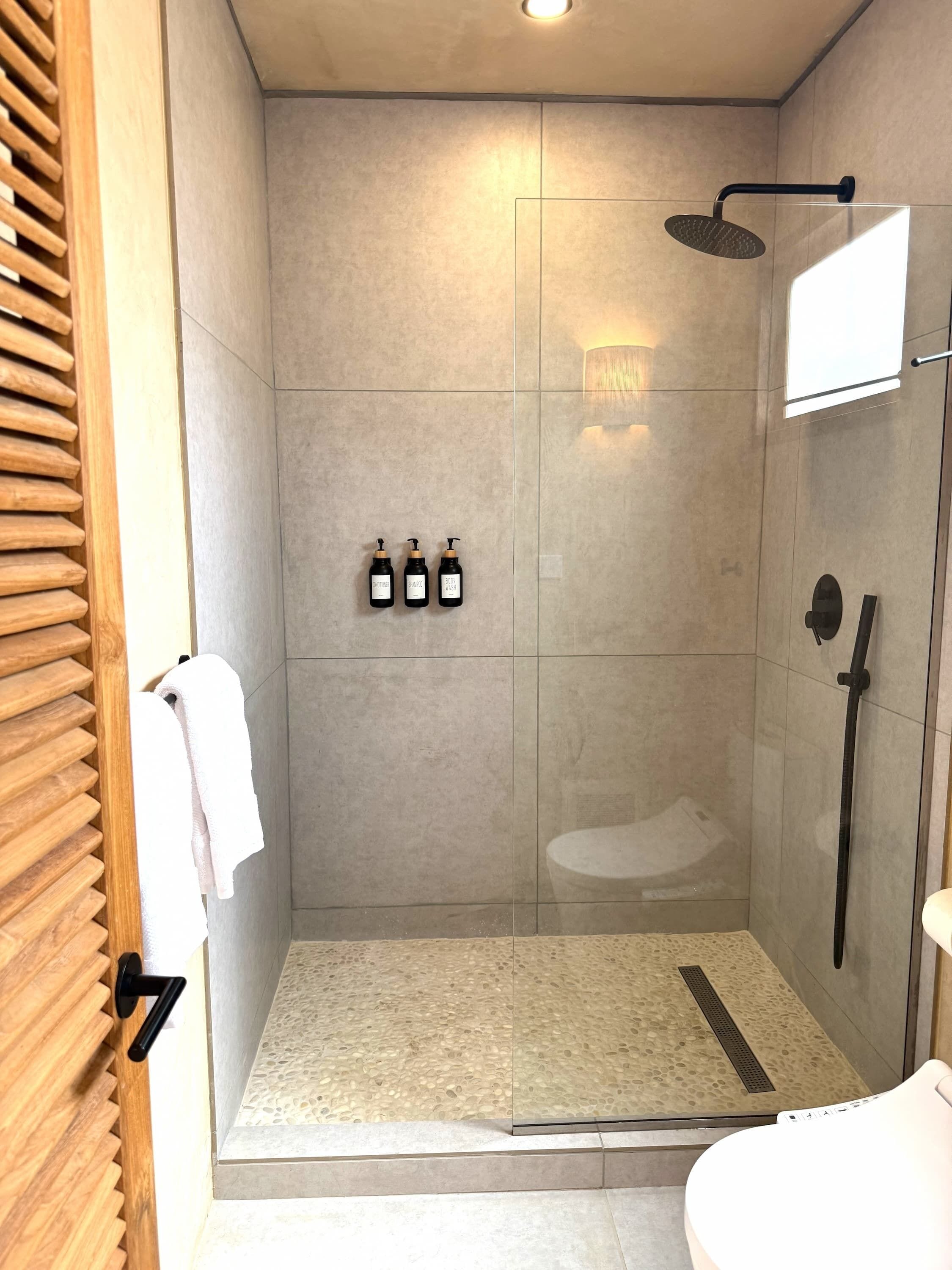 Une douche moderne avec une porte en verre, un pommeau de douche à main et trois bouteilles de shampoing.