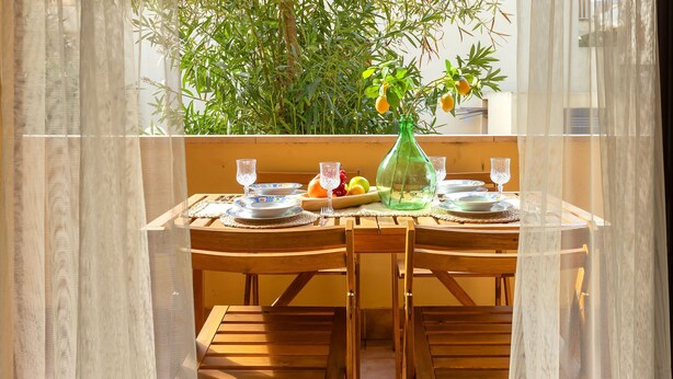 A dining table set with plates, glasses, and a green bottle, surrounded by chairs, with a view of a lush green tree through sheer curtains. - Sardinia Re - Lido 125 (Alghero)