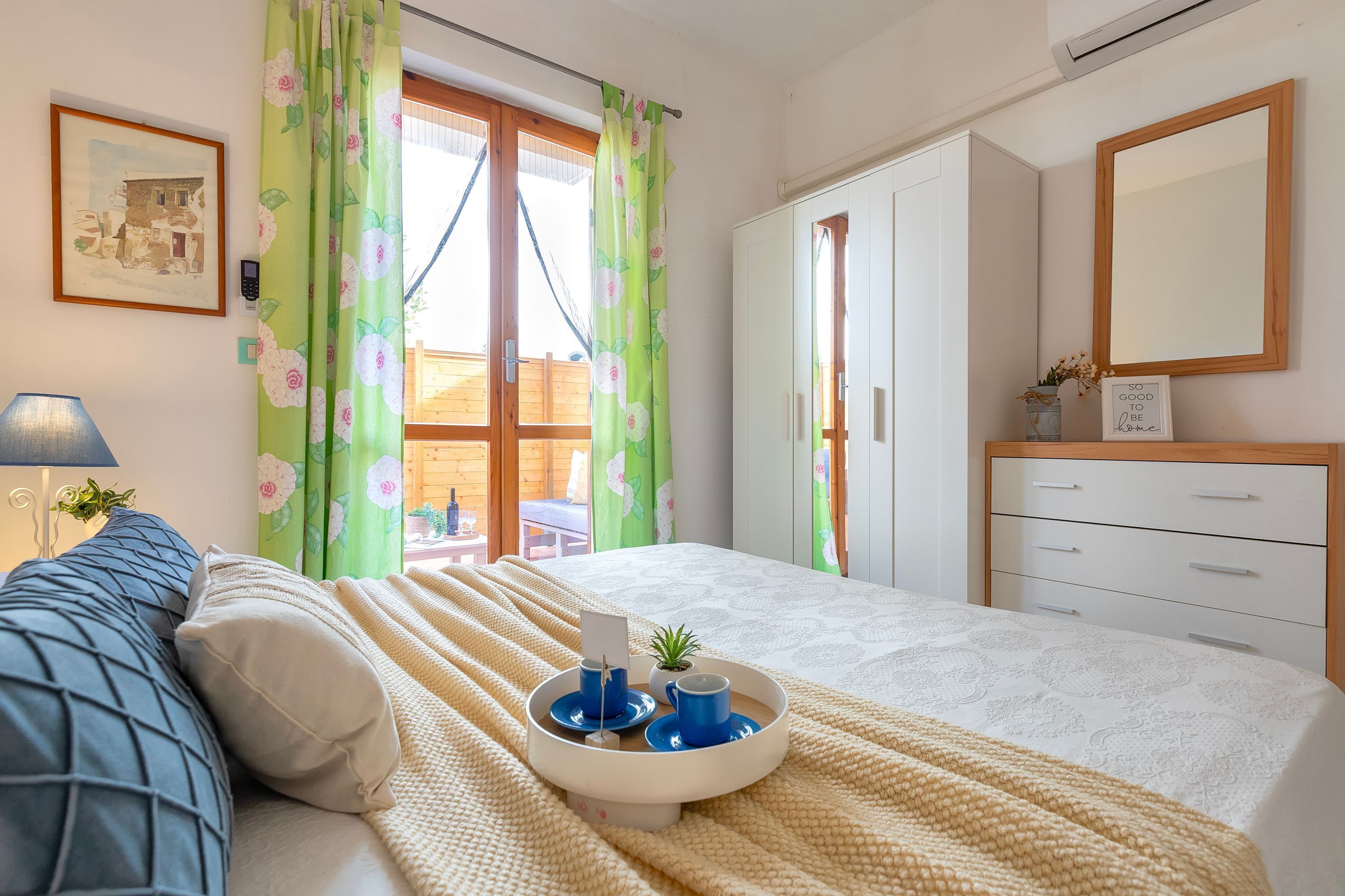 Un dormitorio con una cama, una mesita de noche con una bandeja, una lámpara, un cuadro enmarcado y una ventana con cortinas.