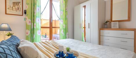 A bedroom with a bed, a bedside table with a tray, a lamp, a framed picture, and a window with curtains.