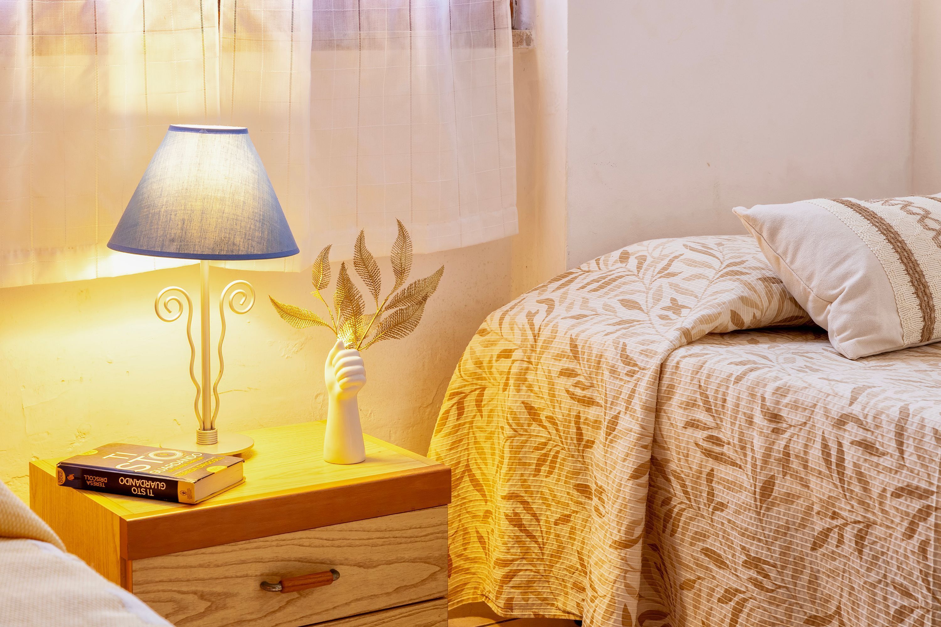 A neatly made bed with a patterned bedspread, a bedside lamp, a book, and a decorative vase on a wooden nightstand.