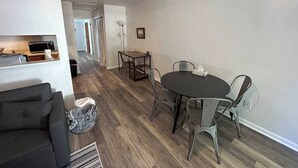 Un salon moderne avec un parquet en bois foncé, un canapé gris et un coin repas comprenant une table ronde et des chaises en métal.