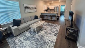 A modern living room with a sofa, coffee table, and a rug. There is a kitchen area with a bar and a ceiling fan.