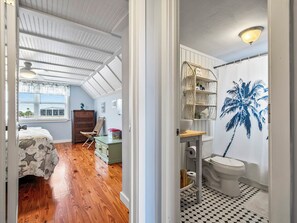 Un dormitorio con piso de madera, una cama con manta de diseño de estrellas, un aparador y una silla. El baño tiene un inodoro blanco, un lavamanos, un espejo y una pared con un mural de palmeras.