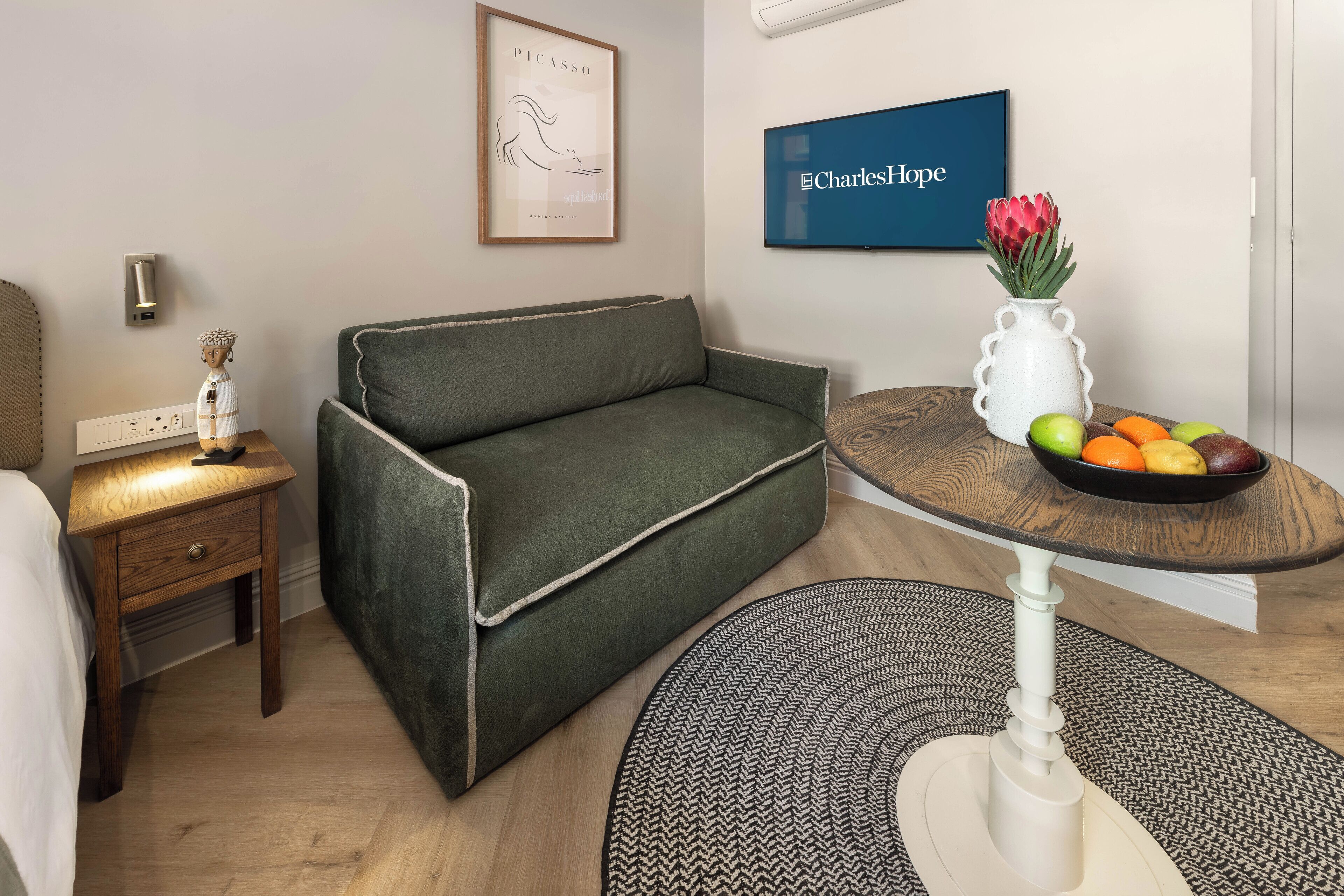 A modern living room with a green sofa, a small wooden side table, a white vase with flowers, and a fruit bowl on a round table.