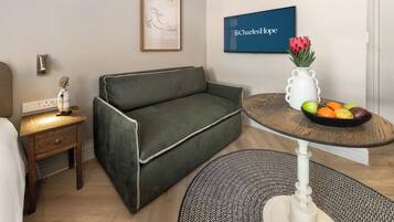 A modern living room with a green sofa, a small wooden side table, a white vase with flowers, and a fruit bowl on a round table.
