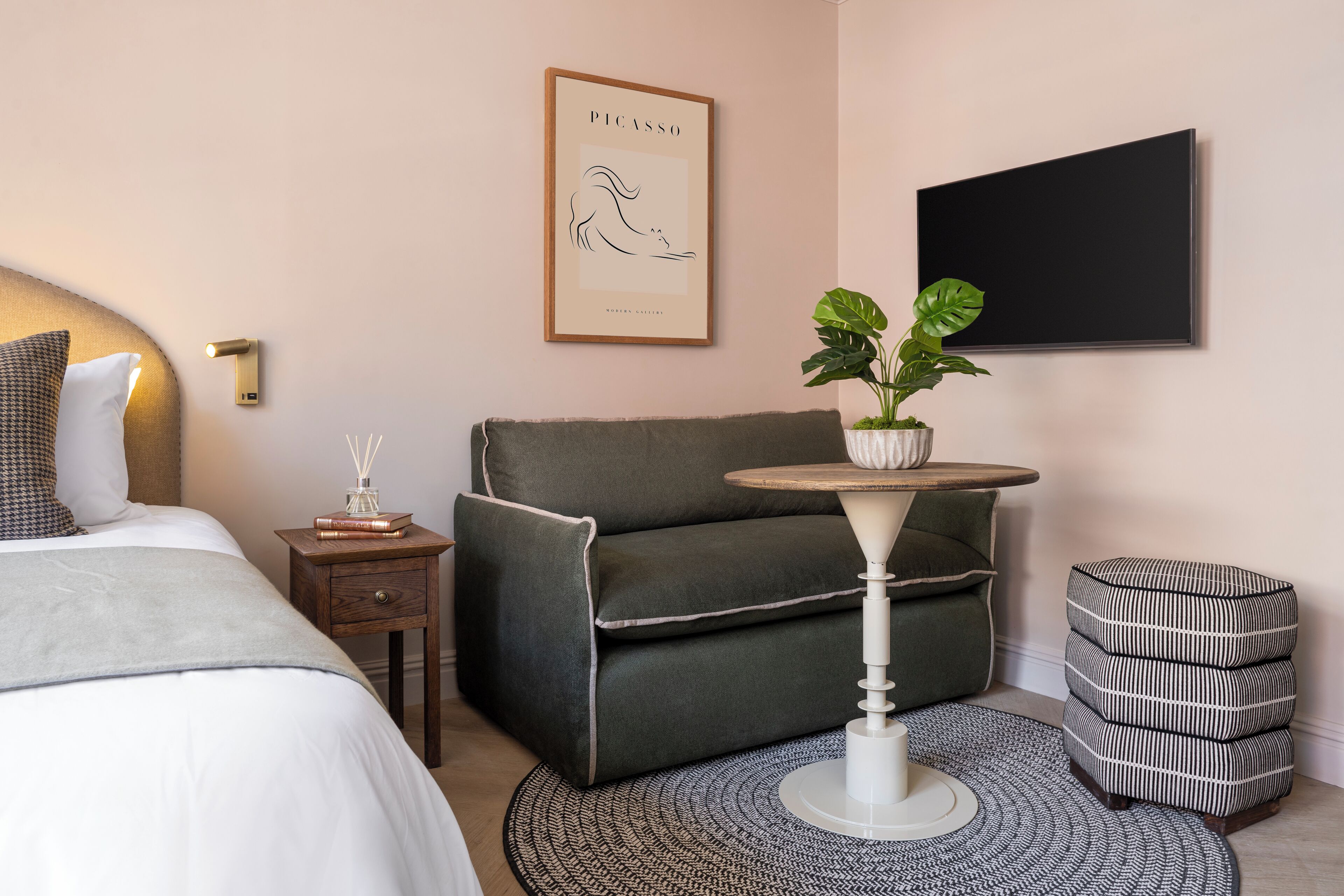 Une chambre d’hôtel moderne avec un lit, un canapé, une petite table agrémentée d’une plante, un tapis rond et une œuvre d’art encadrée au mur.