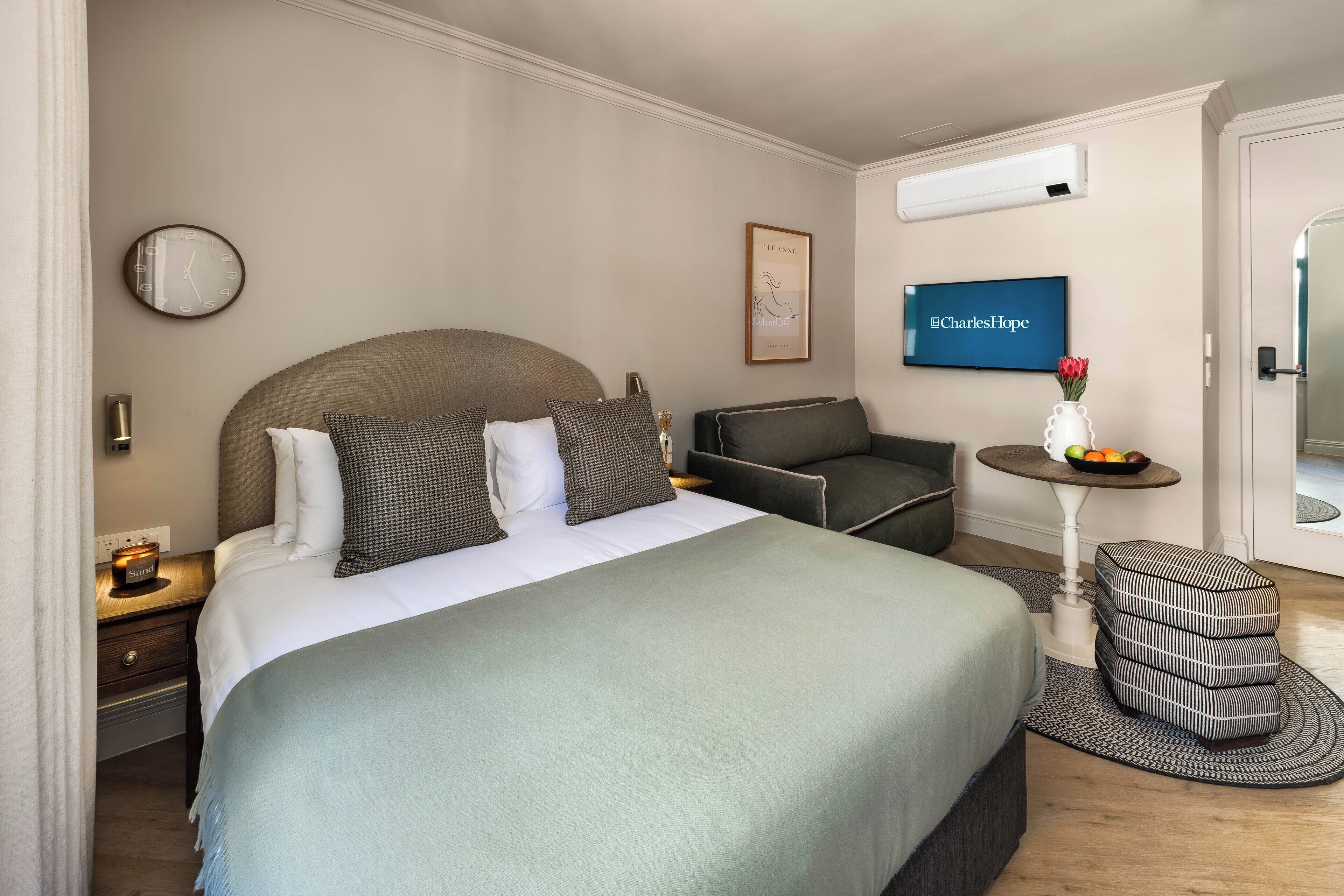 A hotel room with a bed, a chair, a small table with a vase and fruit, a clock, and a mounted television.