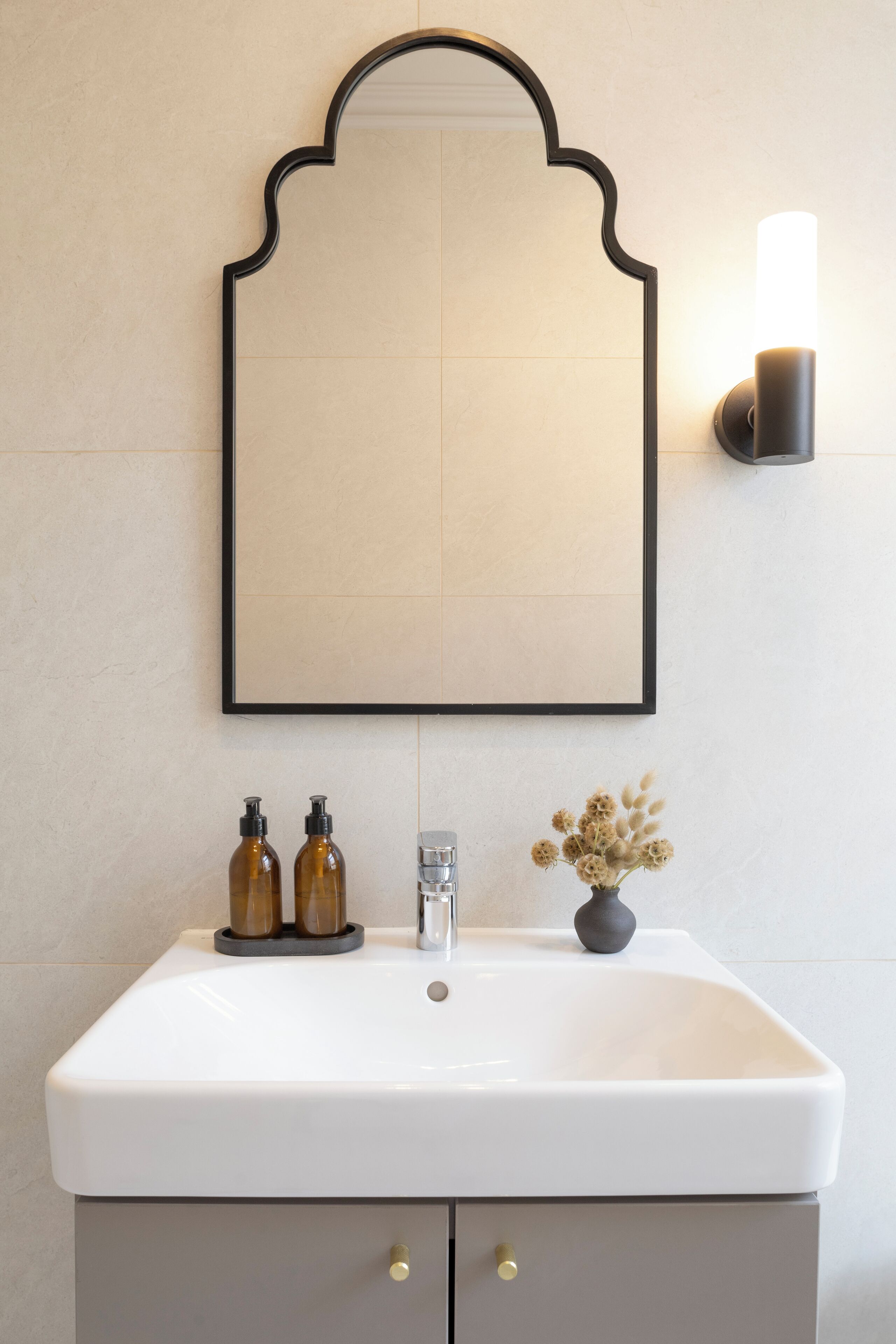 Une salle de bain avec un lavabo blanc, un miroir avec un cadre noir, deux bouteilles marron, un vase avec des fleurs séchées et un luminaire fixé au mur.