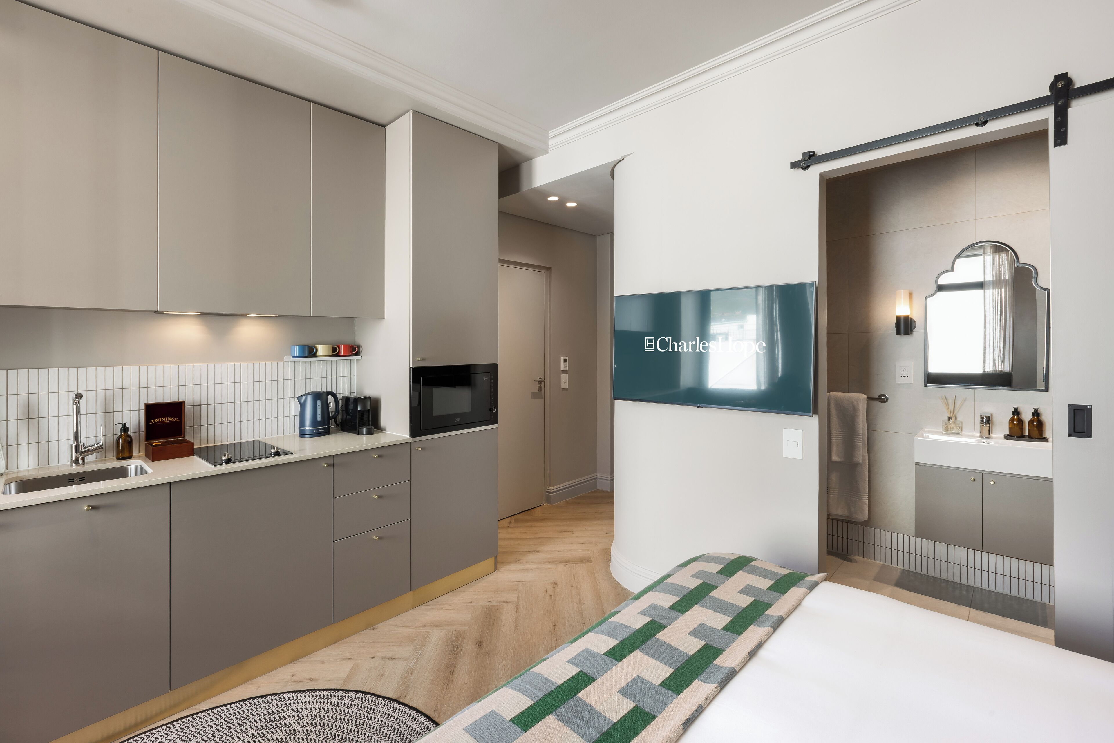 A modern hotel room with a kitchenette, a bed with a patterned bedspread, and a bathroom with a mirror and sink.