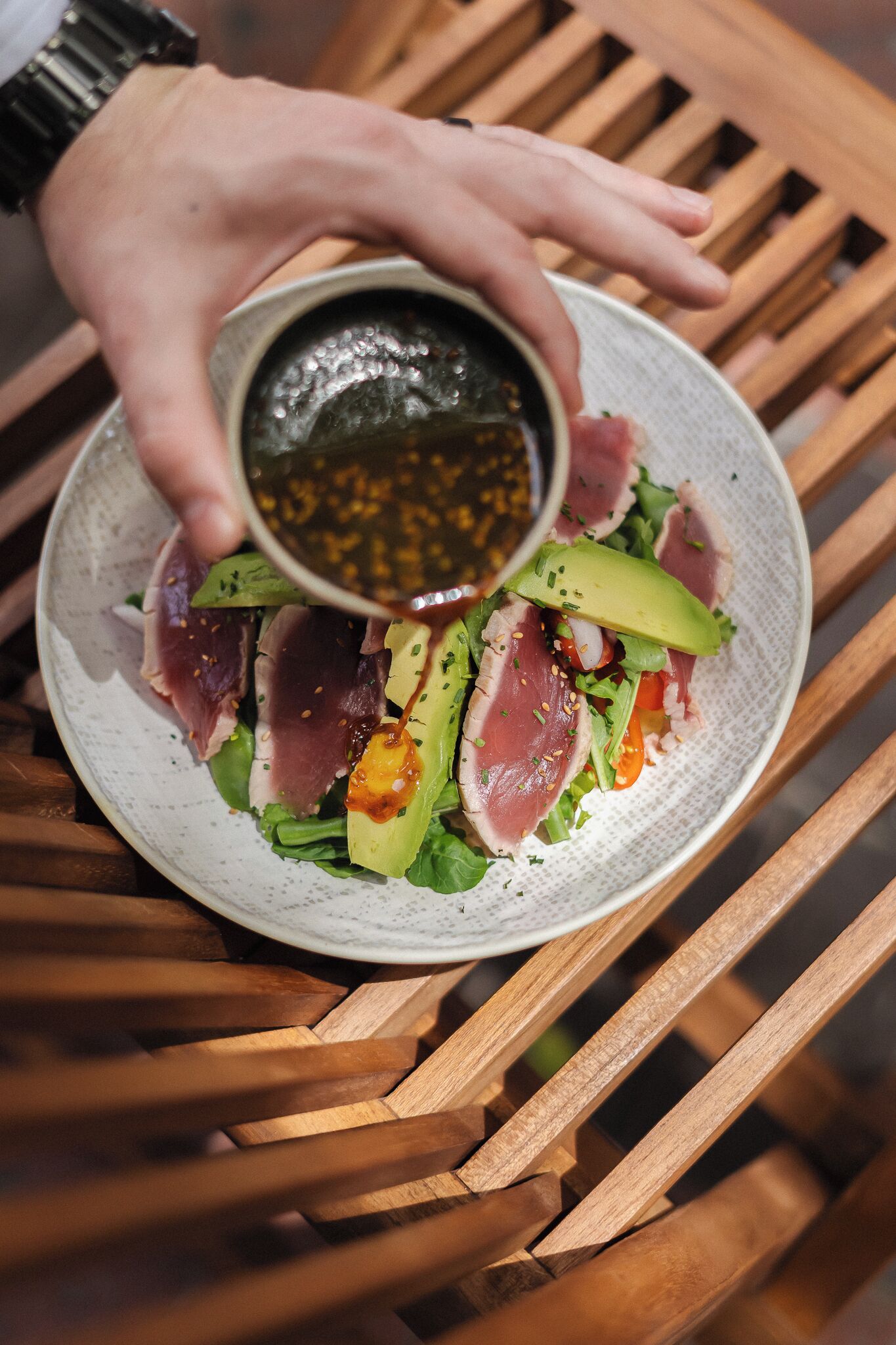 A person pouring sauce over a dish of sliced meat and vegetables on a plate.