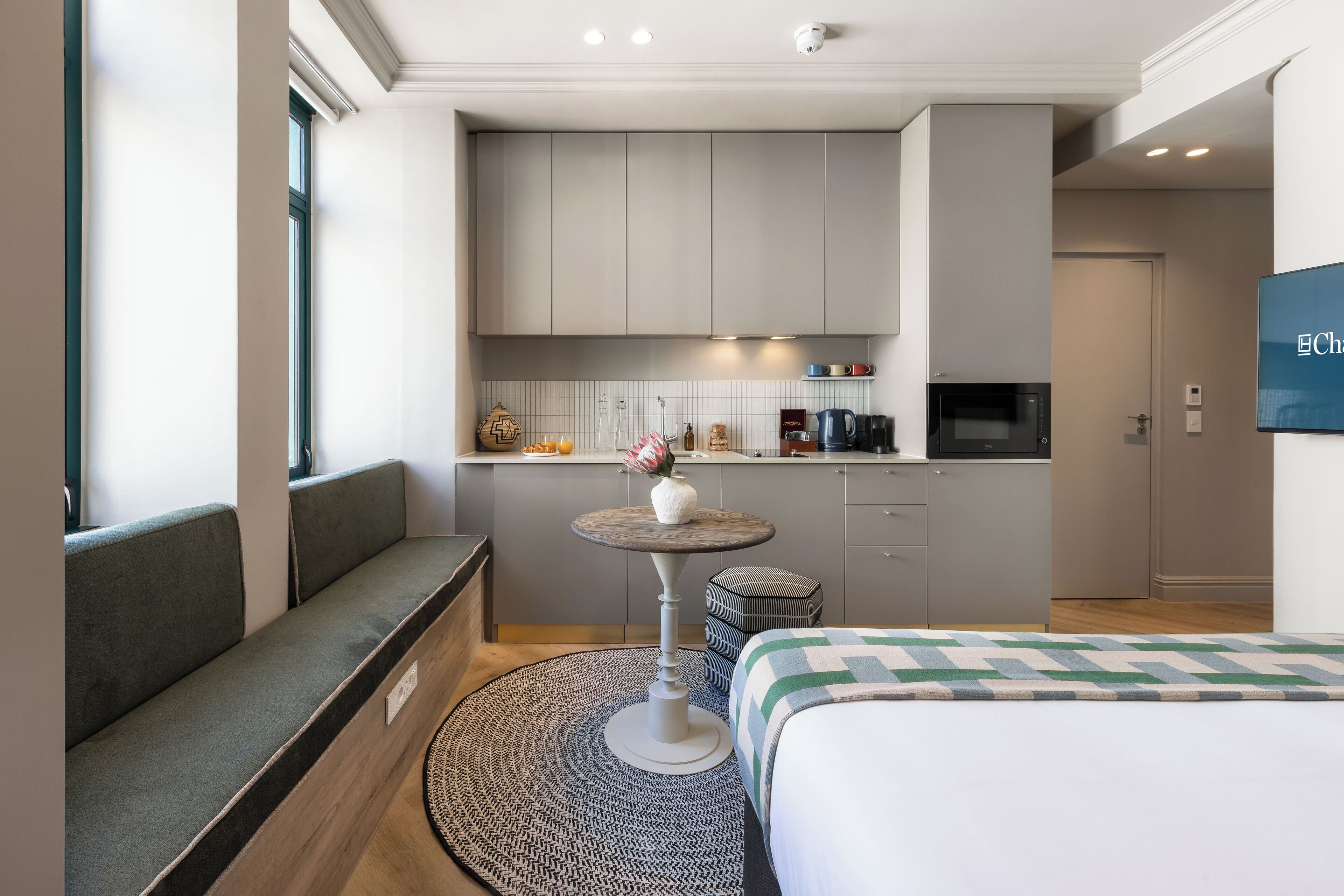 A compact hotel room with a bed, a seating area with a small table, a kitchenette, and a TV mounted on the wall.