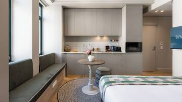 A compact hotel room with a bed, a seating area with a small table, a kitchenette, and a TV mounted on the wall.