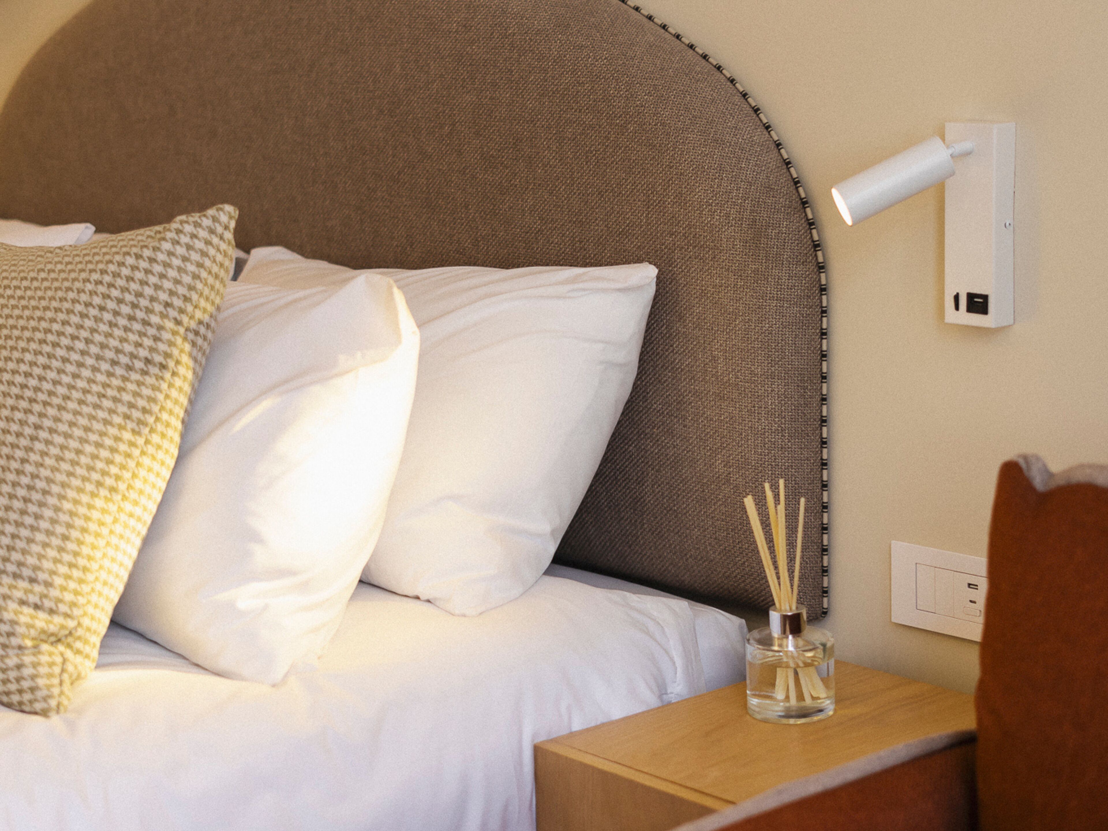 Un lit bien fait, agrémenté d’oreillers blancs et à motifs, une table de chevet avec un diffuseur de parfum à bâtonnets et une lampe fixée au mur.