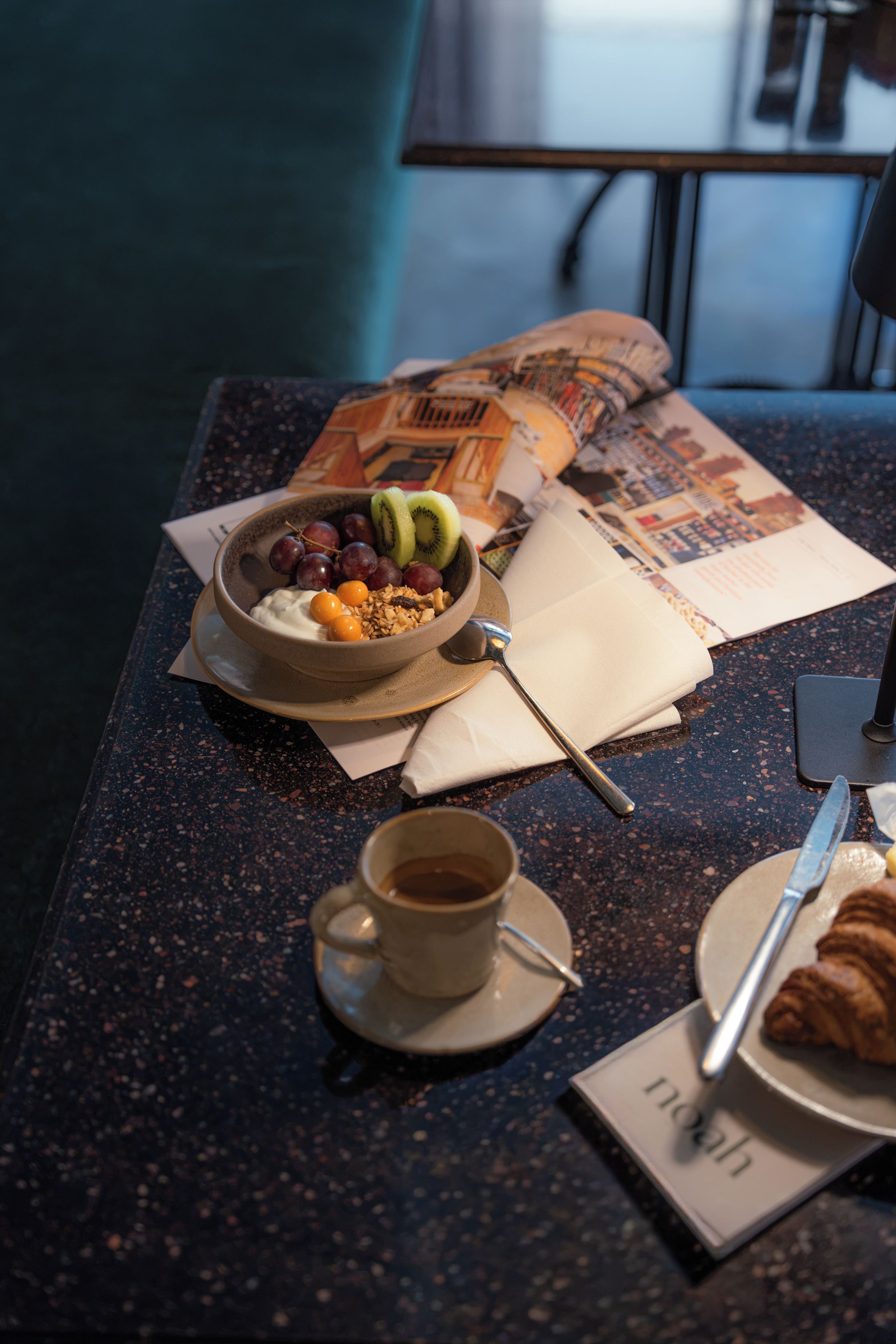 A breakfast setting with a bowl of yogurt and fruits, a cup of coffee, a croissant, and a newspaper on a speckled table.