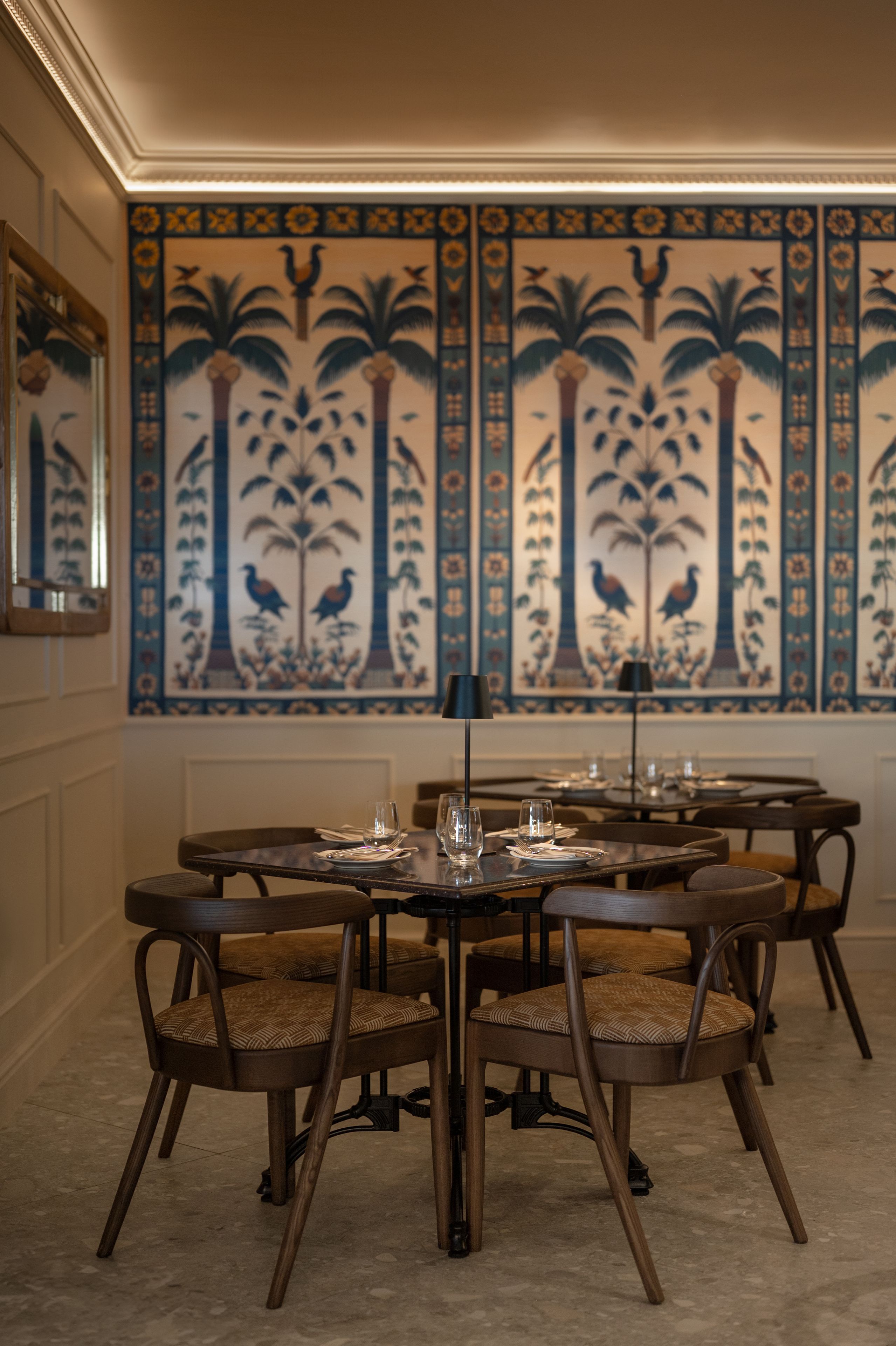 A dining area with round tables, chairs, and a decorative wall with palm trees and birds.