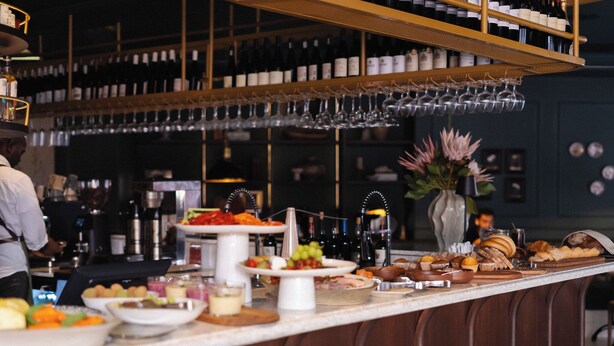 A bar area with a variety of food and drink options, including a buffet with fruits, pastries, and bottles. - Charles Hope Boutique Hometel (Cape Town)