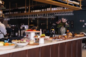 Zona de bar con diversas opciones de comida y bebida, incluyendo un bufé con frutas, pasteles y botellas.