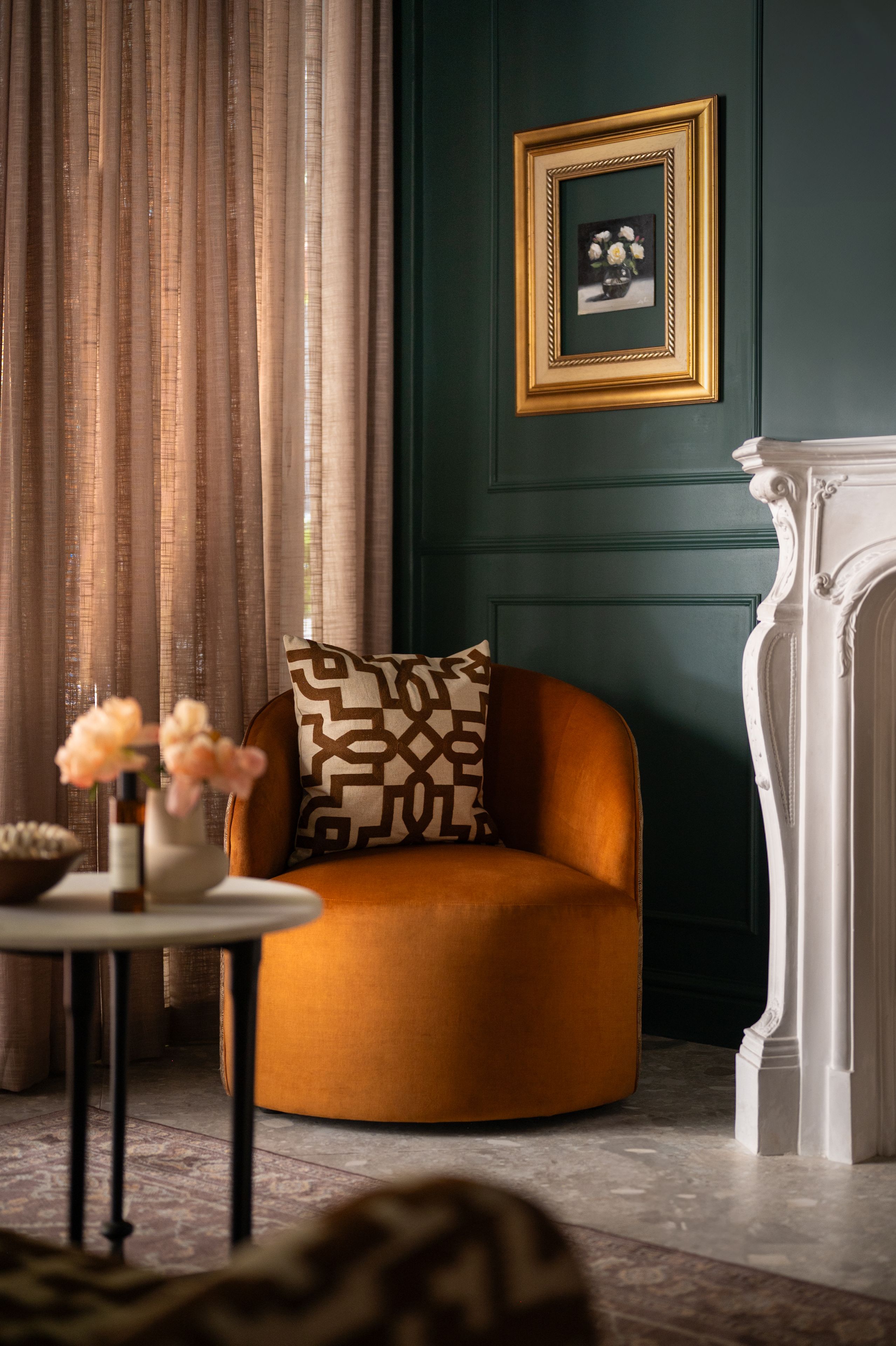 Una habitación con una pared verde oscuro, una chimenea blanca, un sillón marrón con un cojín estampado, una mesita redonda con un jarrón de flores y un cuadro enmarcado en la pared.