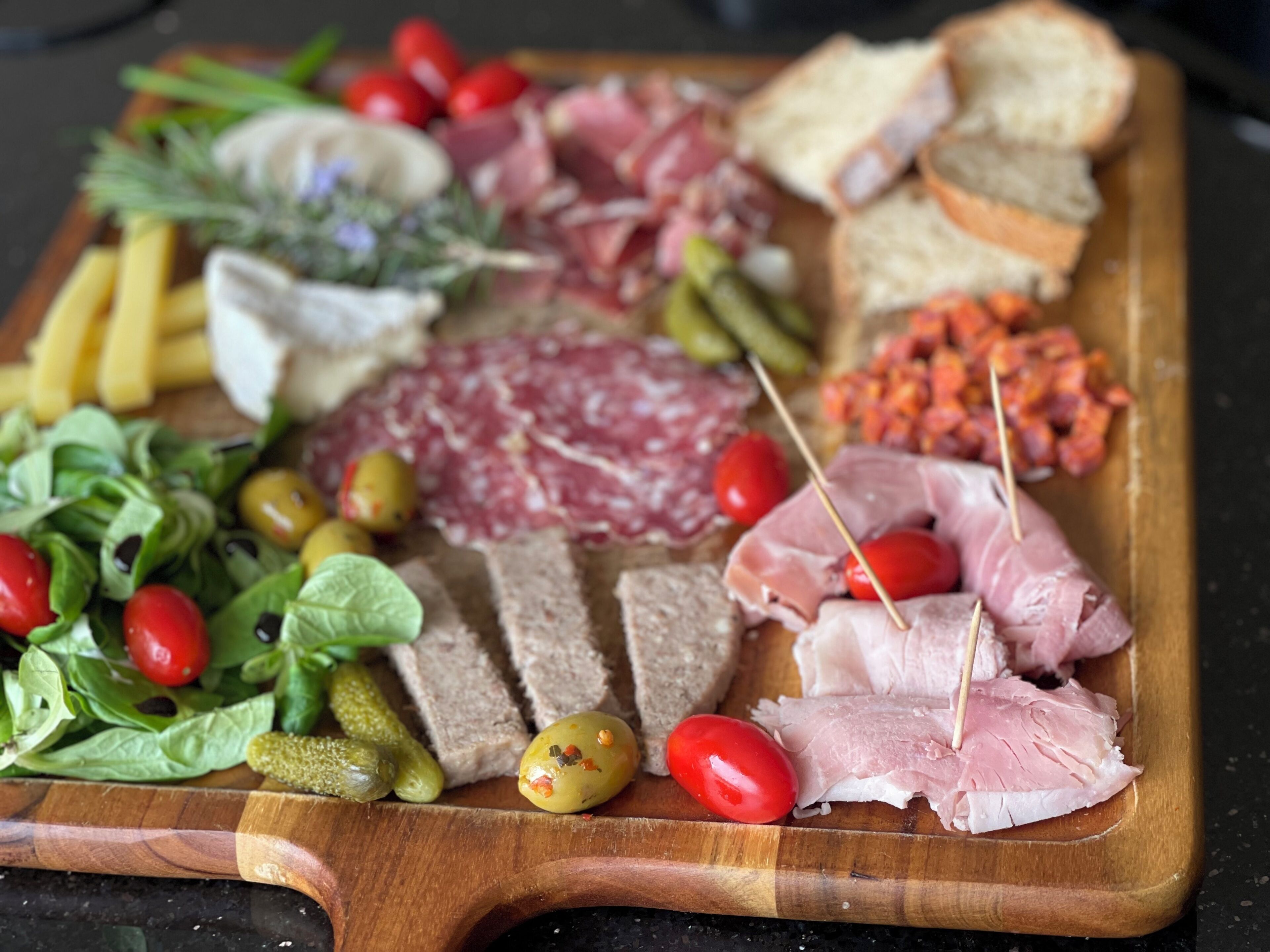 Una tabla de madera con diversos alimentos, como embutidos, pepinillos, aceitunas y pan.