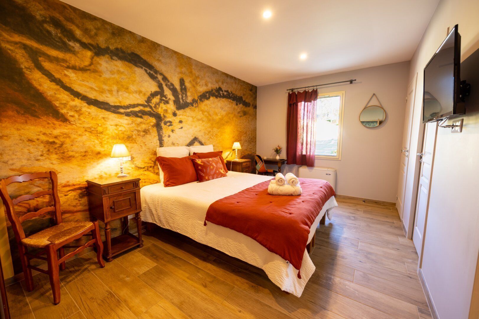 Ein Schlafzimmer mit einem großen Bett, einem Holz-Kopfteil, Nachttischen mit Lampen, einer roten Tagesdecke und einem Wandgemälde.