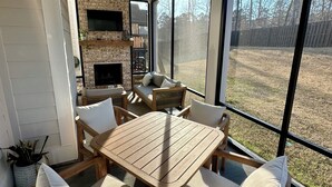 A sunlit patio with a wooden table and chairs, a brick fireplace, and a view of a yard through large windows. - Charming 5-bedroom Masters Rental house in River Oaks  in quaint Evans (Evans)