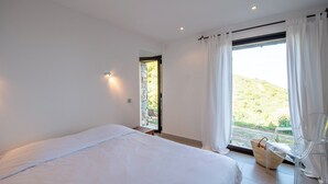 A bedroom with a large bed, a window with curtains, a chair, and a view of a hillside. - Casarone : Havre de paix avec vue sublime sur Calvi, piscine et nature (Calvi)