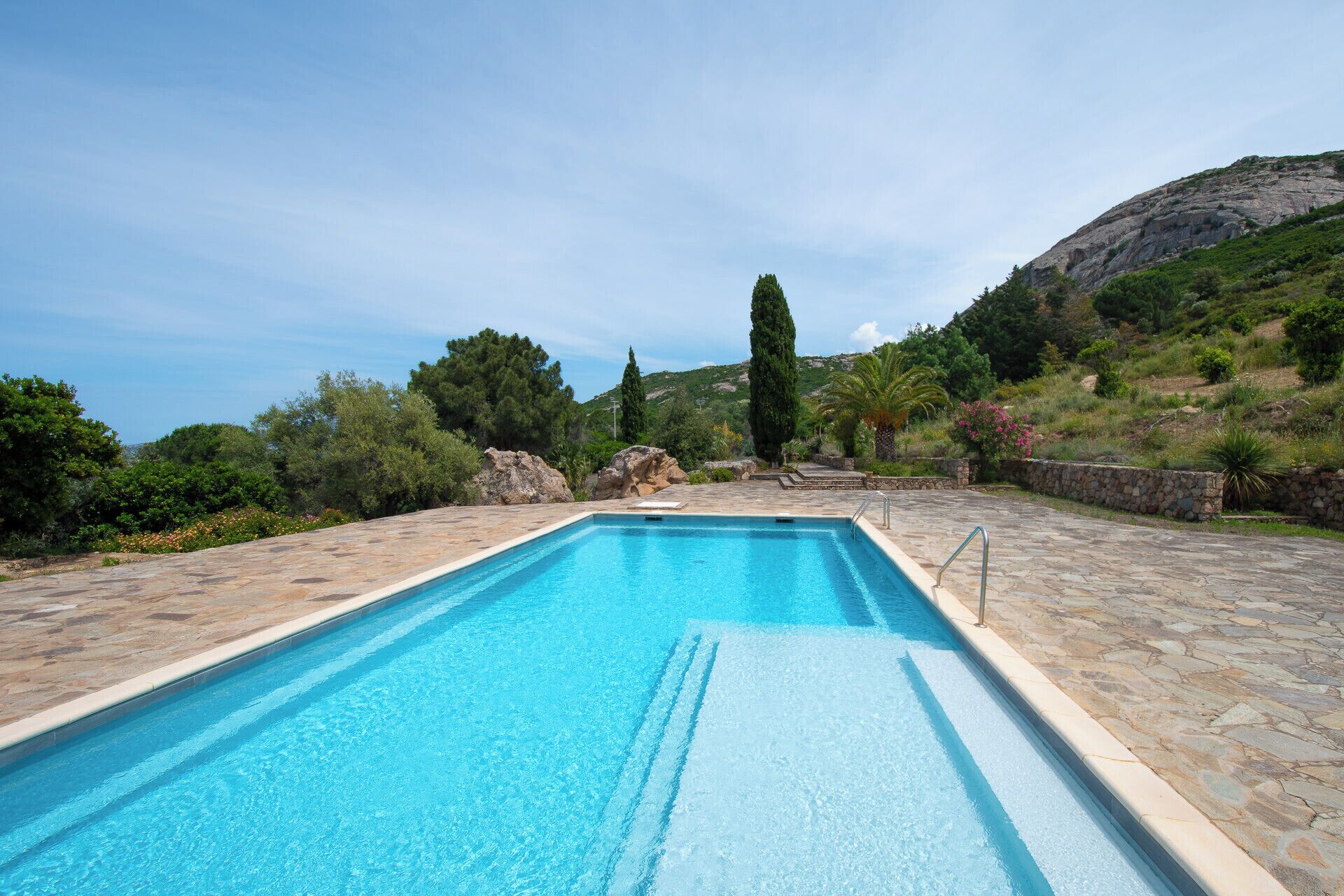 A rectangular swimming pool with clear blue water, surrounded by stone steps and a stone wall.
