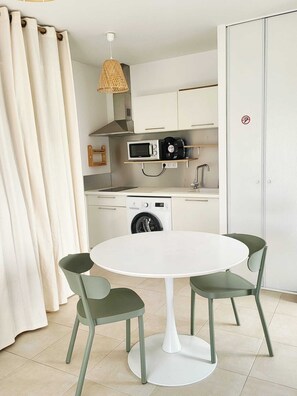 Cocina compacta equipada con microondas, cafetera y lavadora. Hay una mesa redonda con dos sillas y una lámpara colgante.