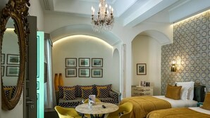 Habitación de hotel con una mesa redonda de comedor, sillas amarillas, un sofá con cojines estampados, una cama con cabecera y un espejo grande.