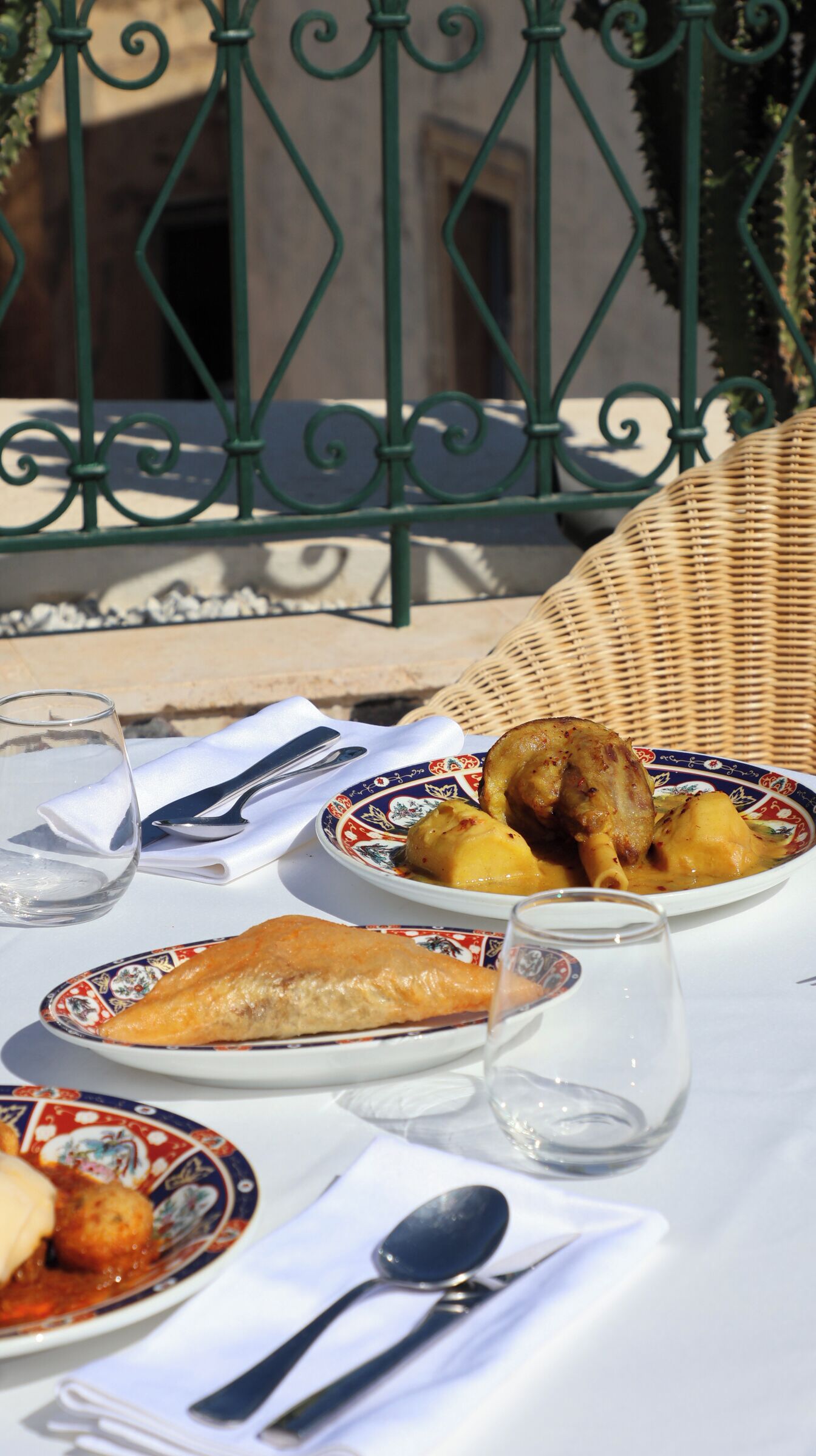 A table set with plates of food, cutlery, and glasses, with a wicker chair and a metal railing in the background.