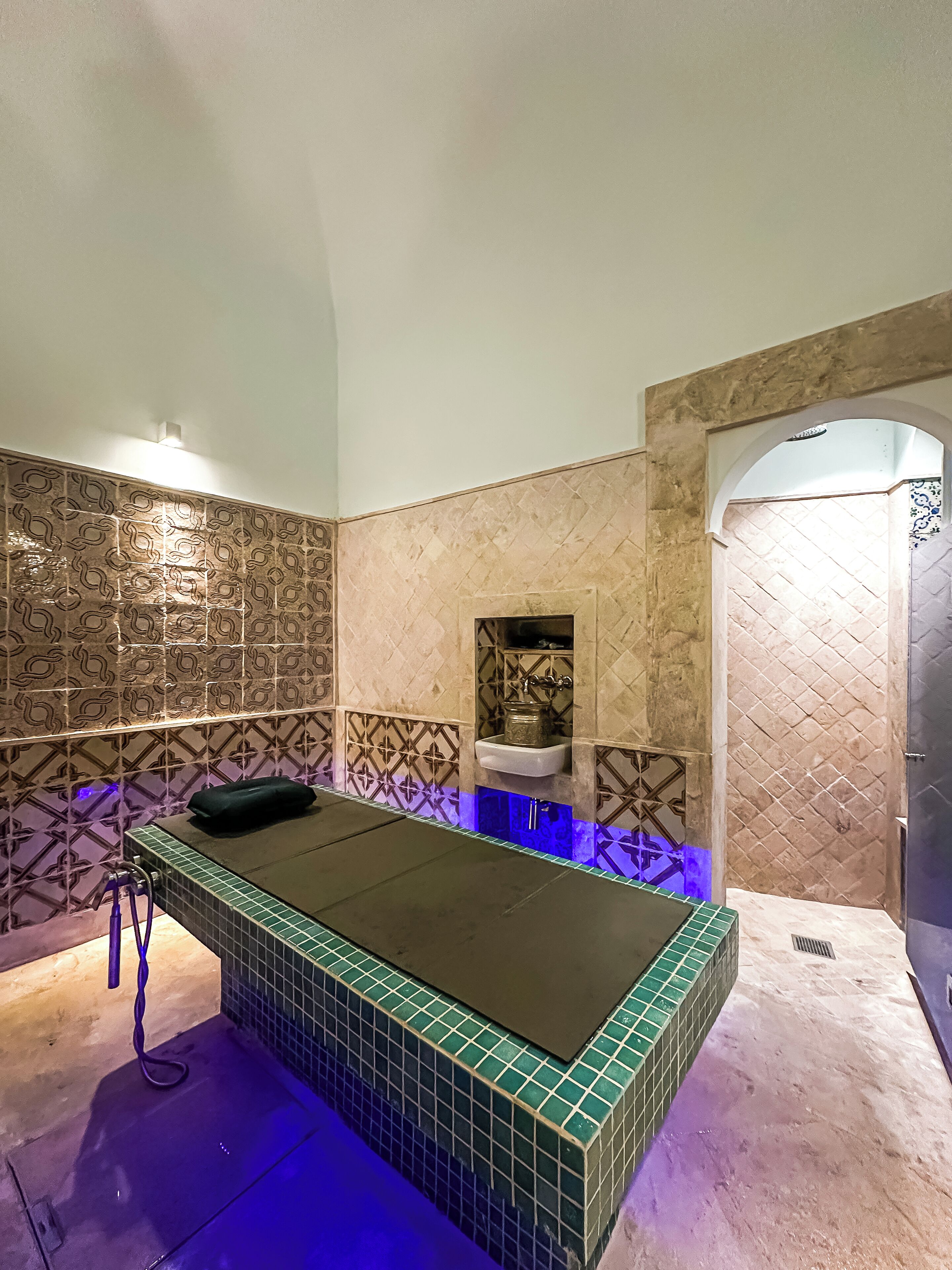 A spa room with a massage table, a sink, and a tiled wall with a geometric pattern.