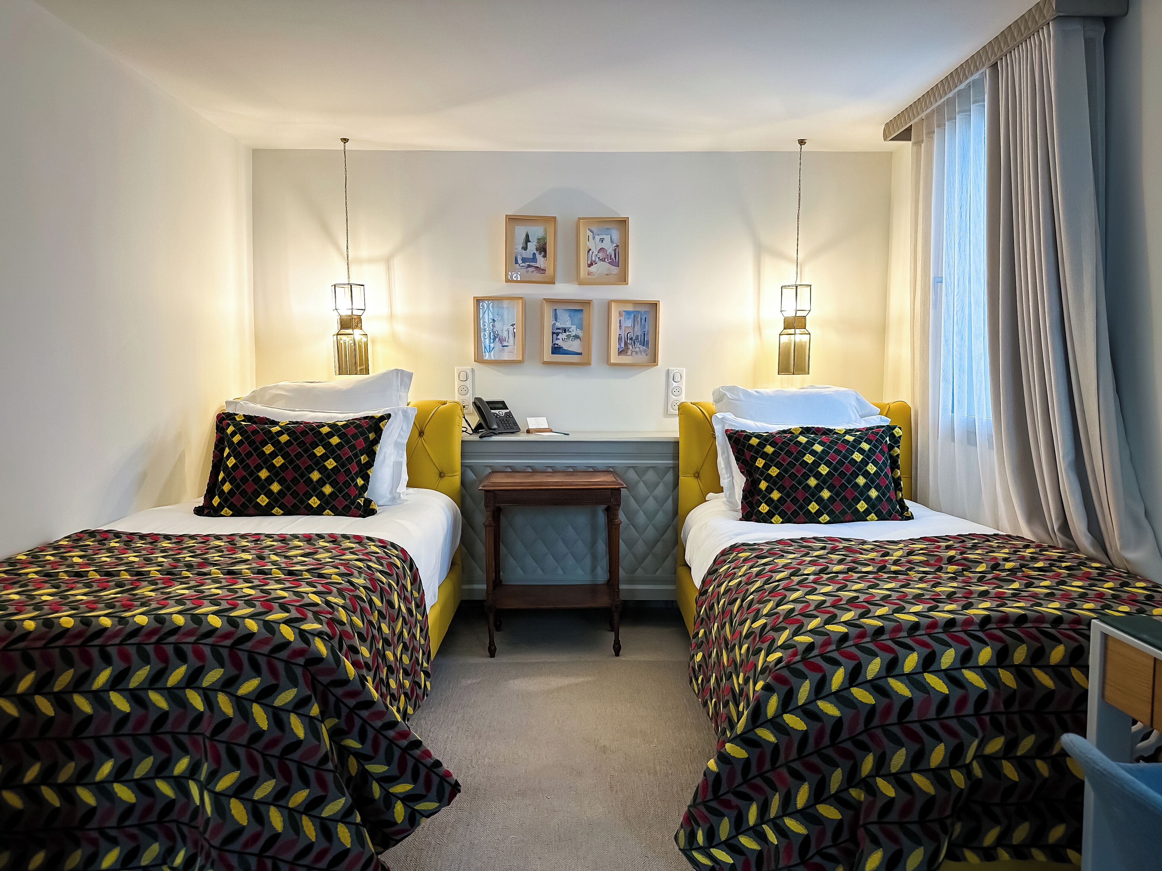 Habitación de hotel con dos camas, un escritorio y una silla. Se observan cuadros enmarcados en la pared, un teléfono y una lámpara.