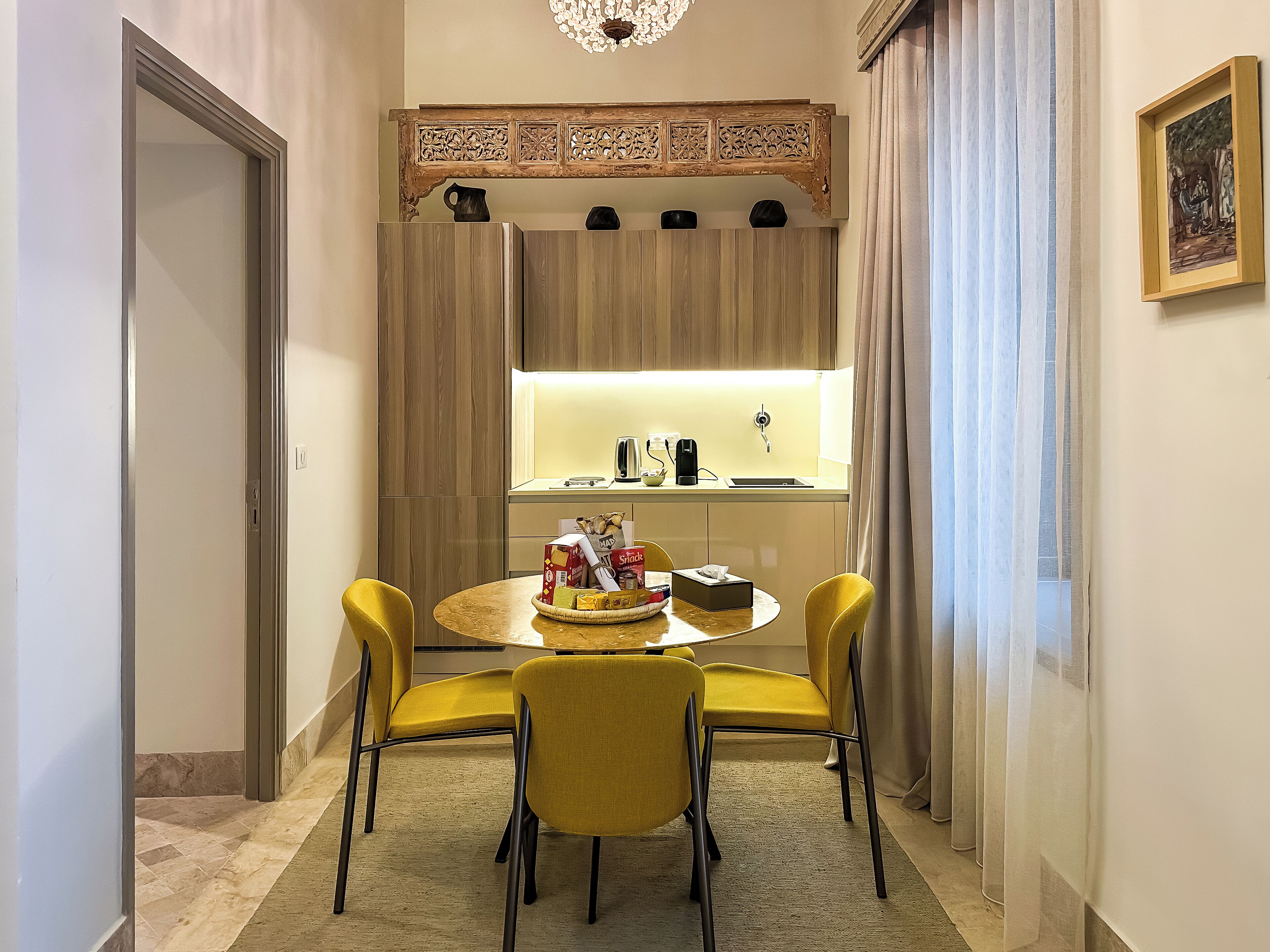 A modern hotel room with a dining area featuring a round table, two yellow chairs, and a decorative wooden cabinet above.