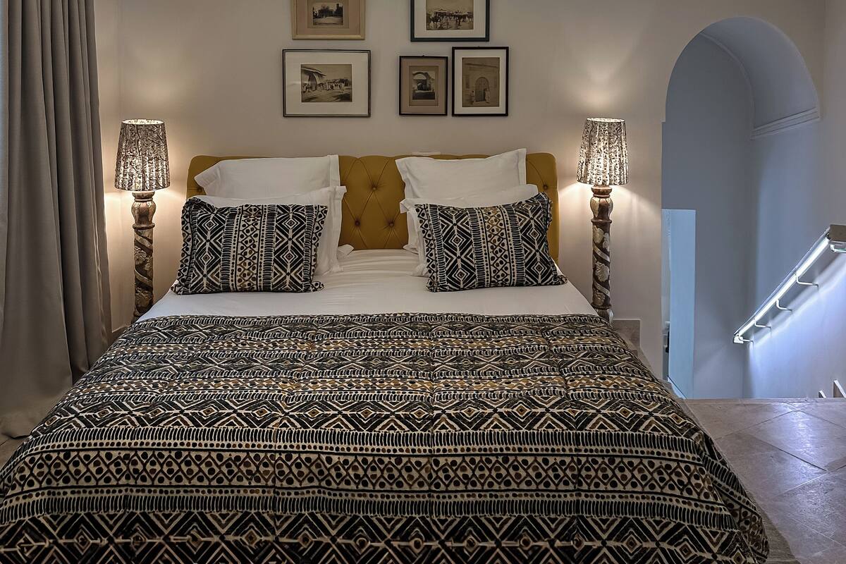 A bedroom with a large bed, patterned bedding, two wall-mounted lamps, and framed pictures on the wall.
