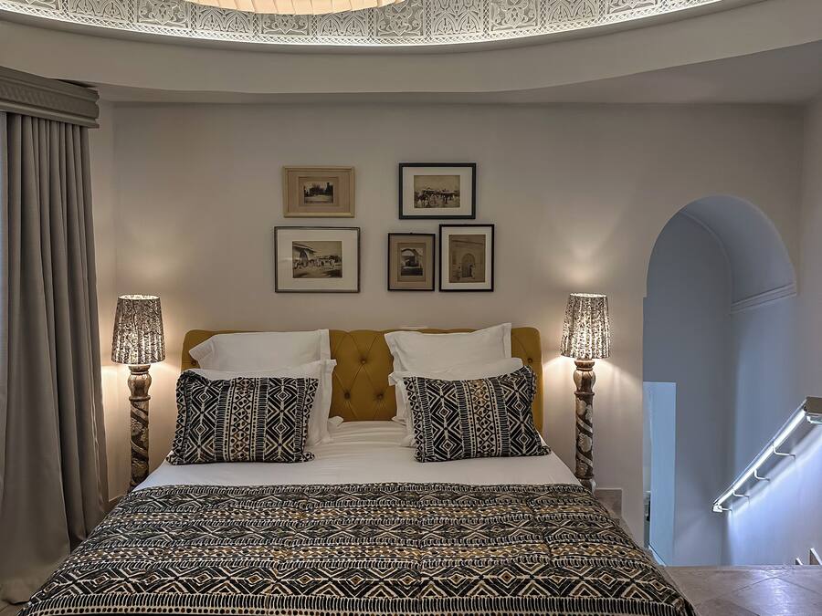Quarto com uma cama grande, roupa de cama estampada, duas luminárias fixadas na parede e quadros enquadrados.
