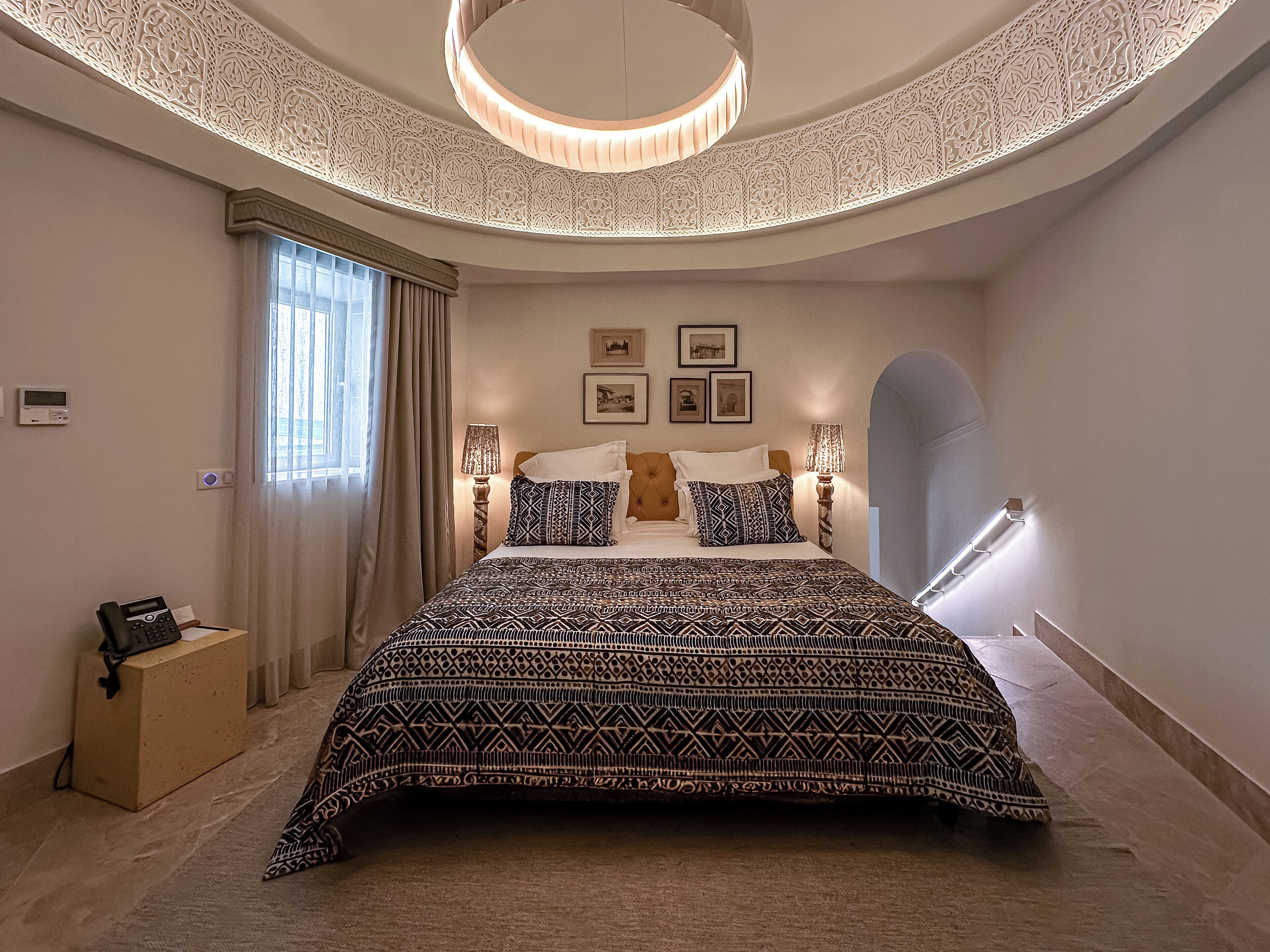 A modern hotel room with a large bed, a round ceiling light, and a view of the city through the window.