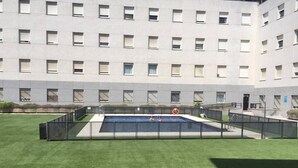 Ein Schwimmbecken mit zwei Personen im Wasser, umgeben von einem Zaun, vor einem Gebäude mit vielen Fenstern.